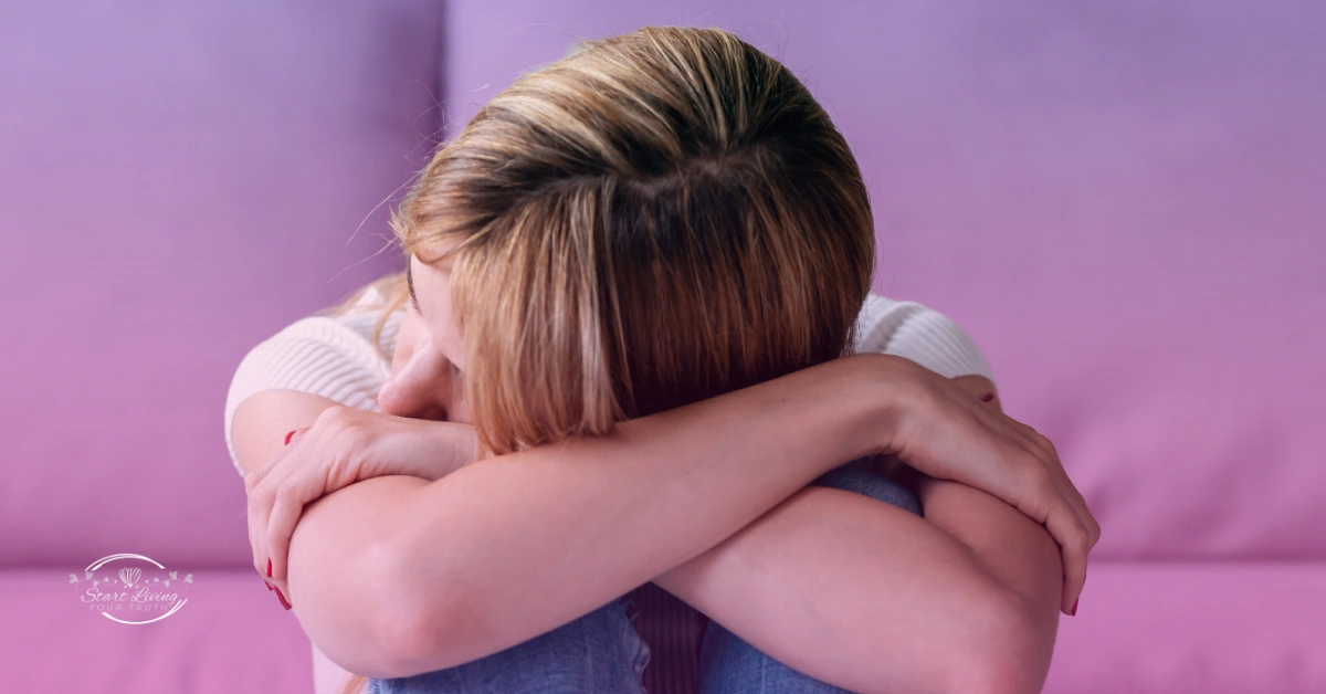 Person sitting with arms around knees, looking down.